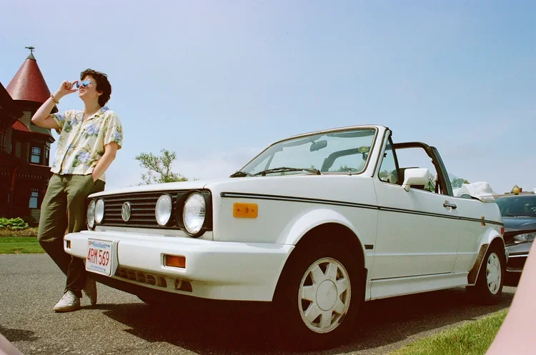 A woman standing next to a white vintage Volkswagen convertible car on a sunny day.