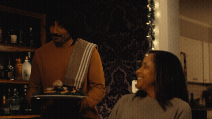 A man serving a woman at an indoor gathering or restaurant, with a dark patterned wall and shelves with bottles in the background.