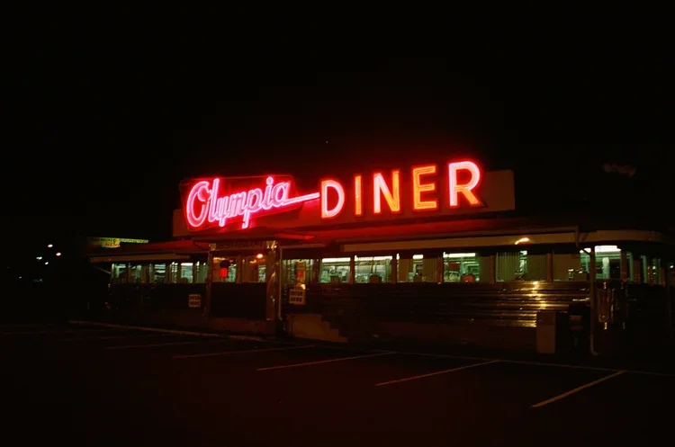 Night view of Olympia Diner with neon sign, featuring a vintage bus in front and empty parking lot.