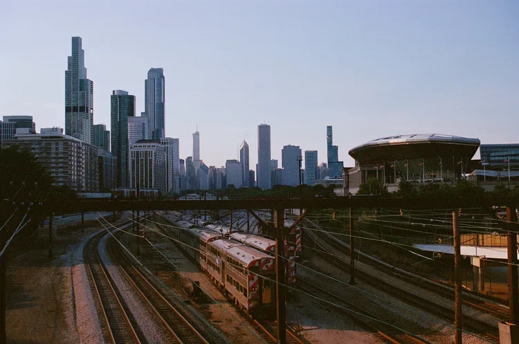 A city skyline with tall skyscrapers in the background, train tracks with a passenger train in the foreground, and a stadium-like structure on the right.