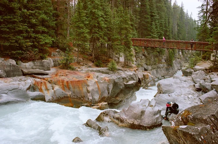 A person taking photographs by a river with rocks, trees, and a wooden bridge in the background.