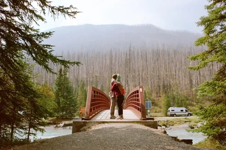 A family on a small wooden bridge over a river in a forested area with mountains in the background.