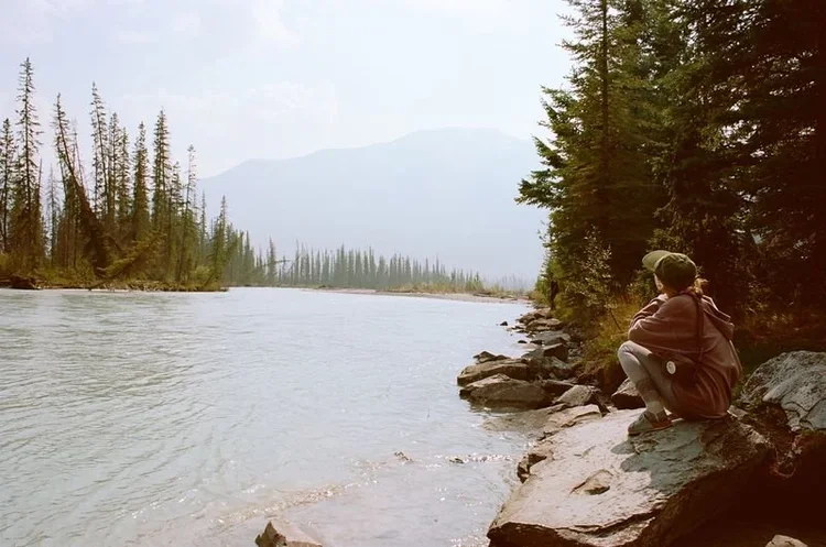 Person sitting on rocks by a river in a forested area, with mountains in the distance, during daytime.