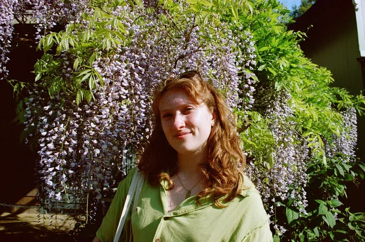 A young woman with red hair stands in front of blooming wisteria flowers and green foliage in a garden during daytime.
