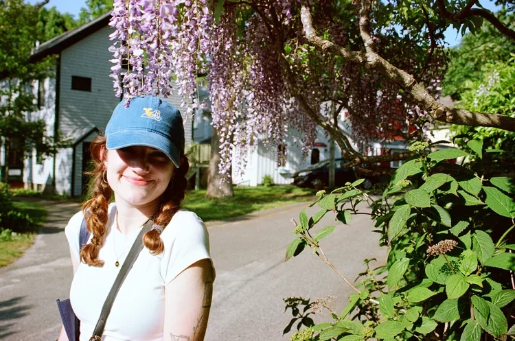 A smiling woman with red hair in braids wearing a blue cap, standing outdoors near a flowering tree with purple blossoms in a suburban neighborhood.