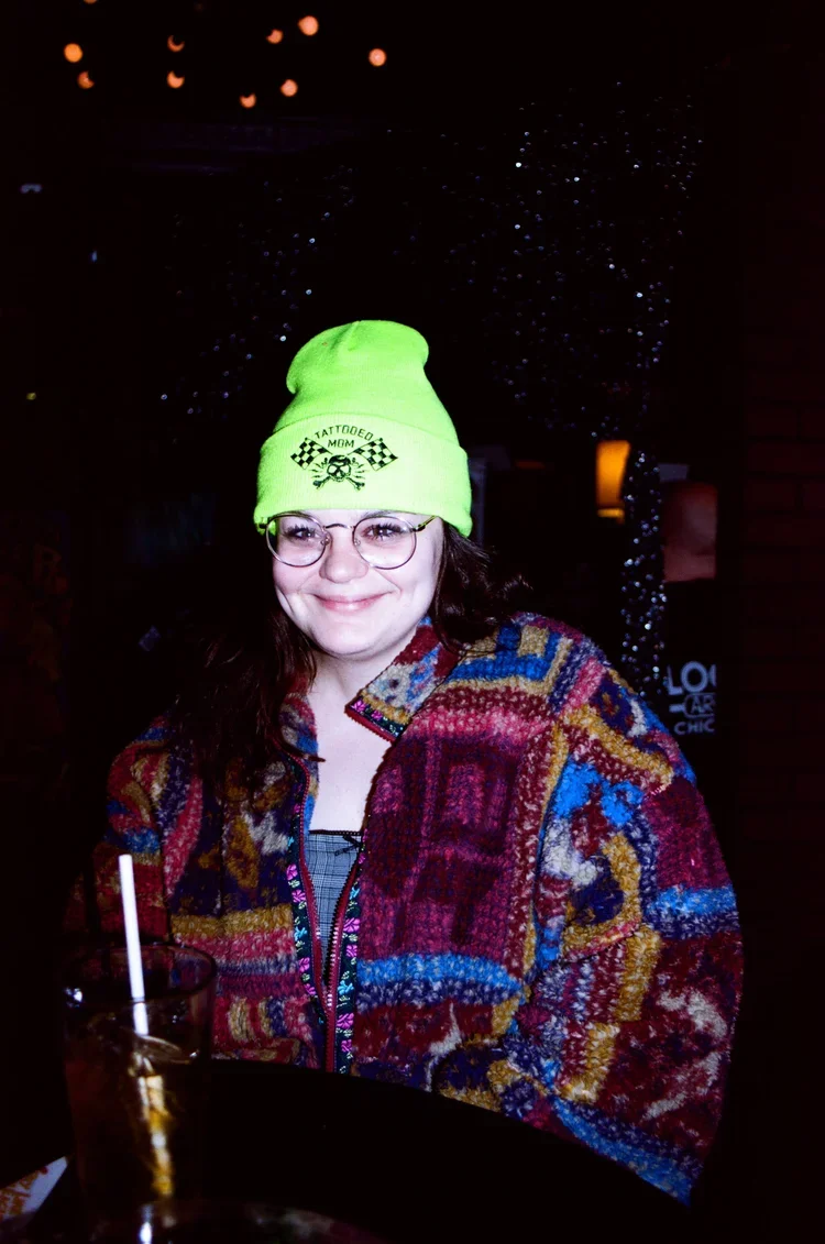 A woman smiling at a bar, wearing round glasses, a bright green beanie with a skull and checkered flag design, and a colorful patchwork jacket. There is a drink with a straw on the bar in front of her. The background is dark with small light dots and