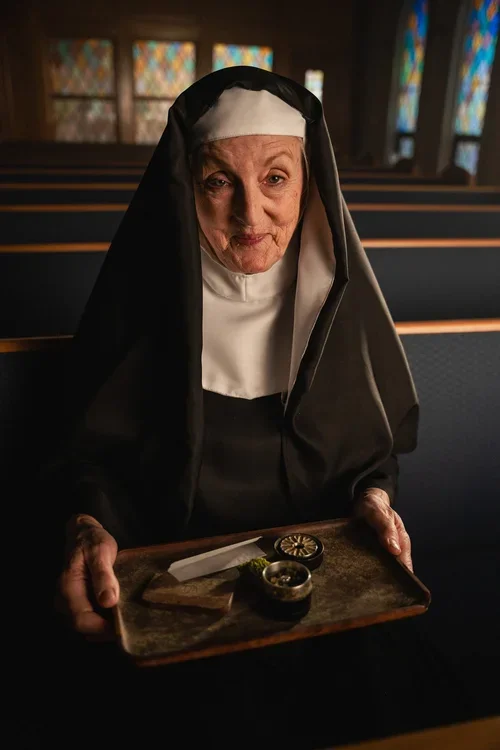 An elderly nun holding a wooden tray with a small bowl of incense, a lit candle, and a piece of paper inside a church with stained glass windows.