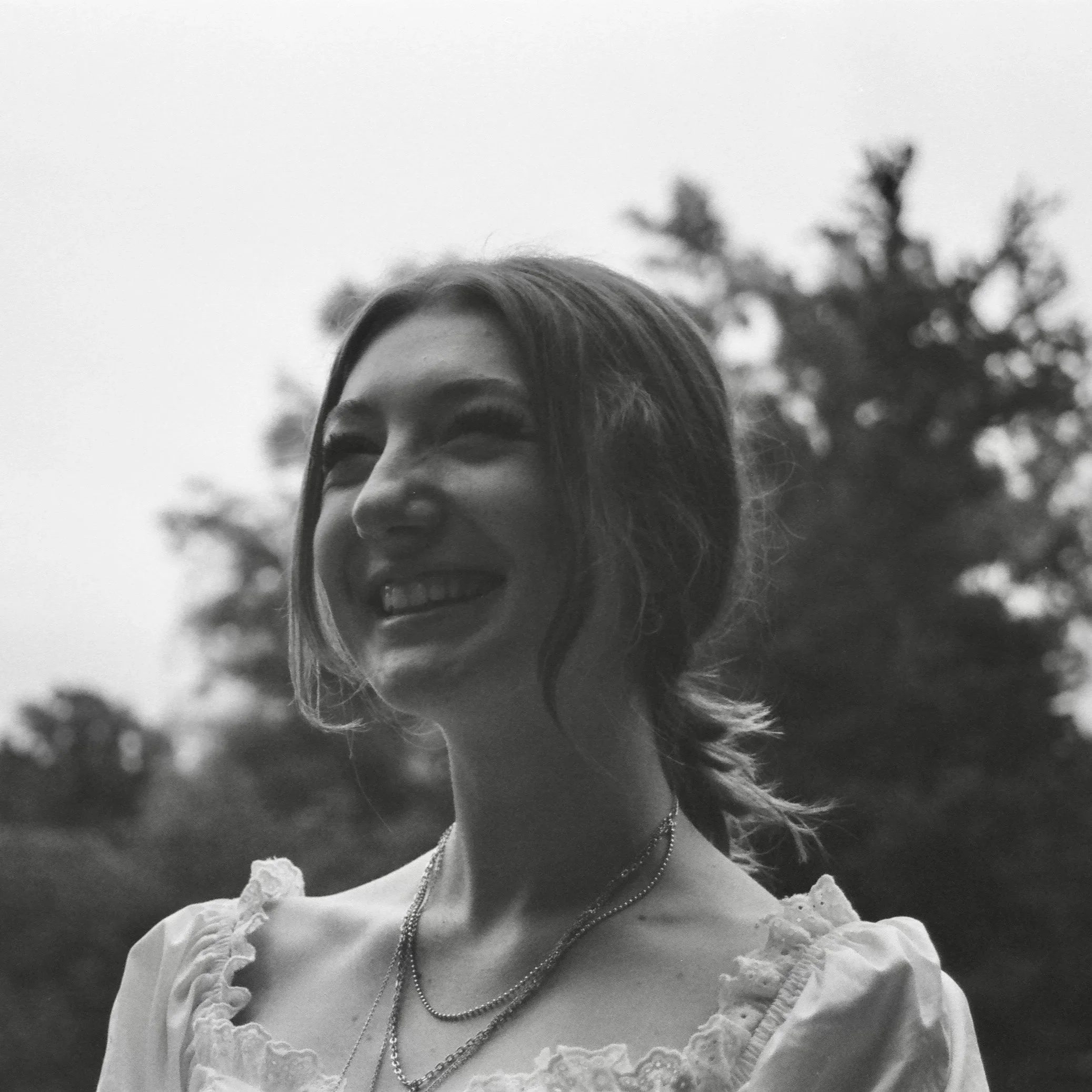 A black and white photo of a woman with a big smile, wearing multiple necklaces, a top with ruffled or lace details, and sunglasses. Trees are visible in the background.