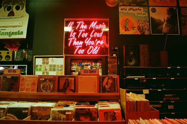A record store interior with vinyl records on display, a neon sign reading 'The music is too loud then you're too old', and posters on the wall.