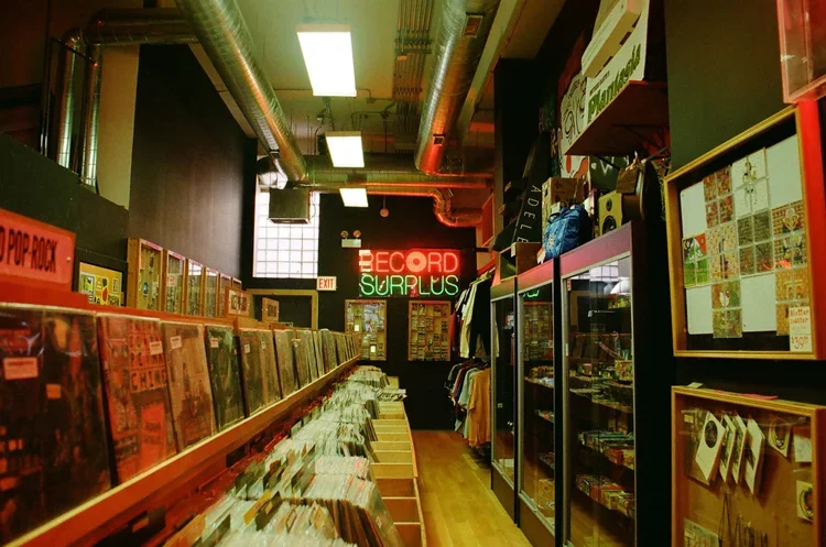 Interior of a record store with vinyl records on shelves, display cases, and a neon sign that reads 'RECORD SURPLUS.'