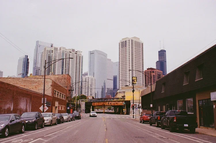 City street with parked cars, brick and dark-colored buildings, and a skyline of tall skyscrapers in the background.