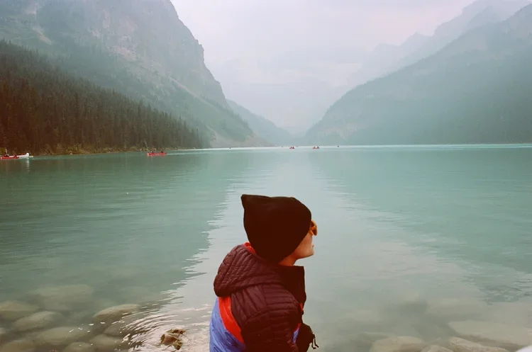 Child sitting by a calm lake surrounded by mountains and forest, with a misty sky overhead.