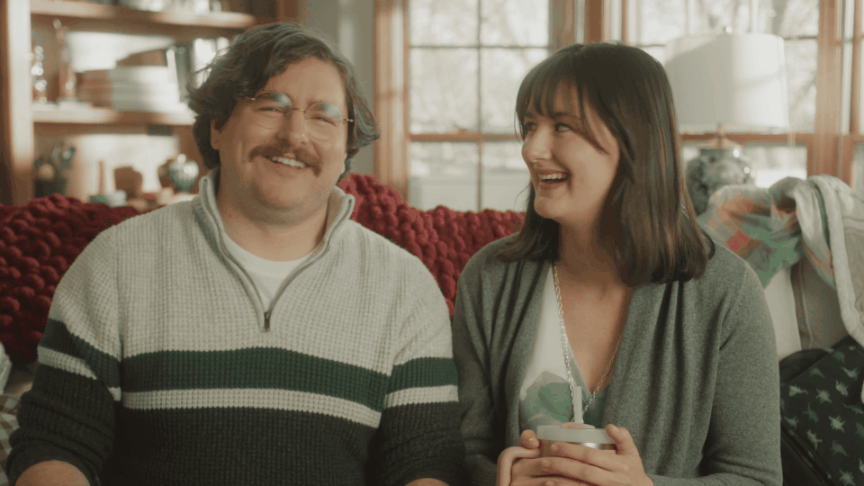 A man with glasses and a mustache, and a woman with dark hair, sitting on a cozy couch, smiling and laughing while holding a mug in a warmly lit room with large windows and wooden frames.