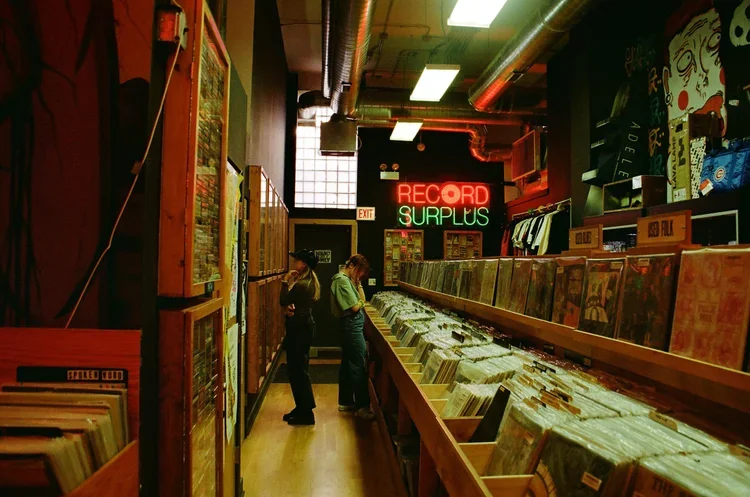 Interior of a record store with wooden shelves filled with vinyl records and a neon sign that says 'RECORD SURPLUS' in red and green. Two people are browsing the records in the background.