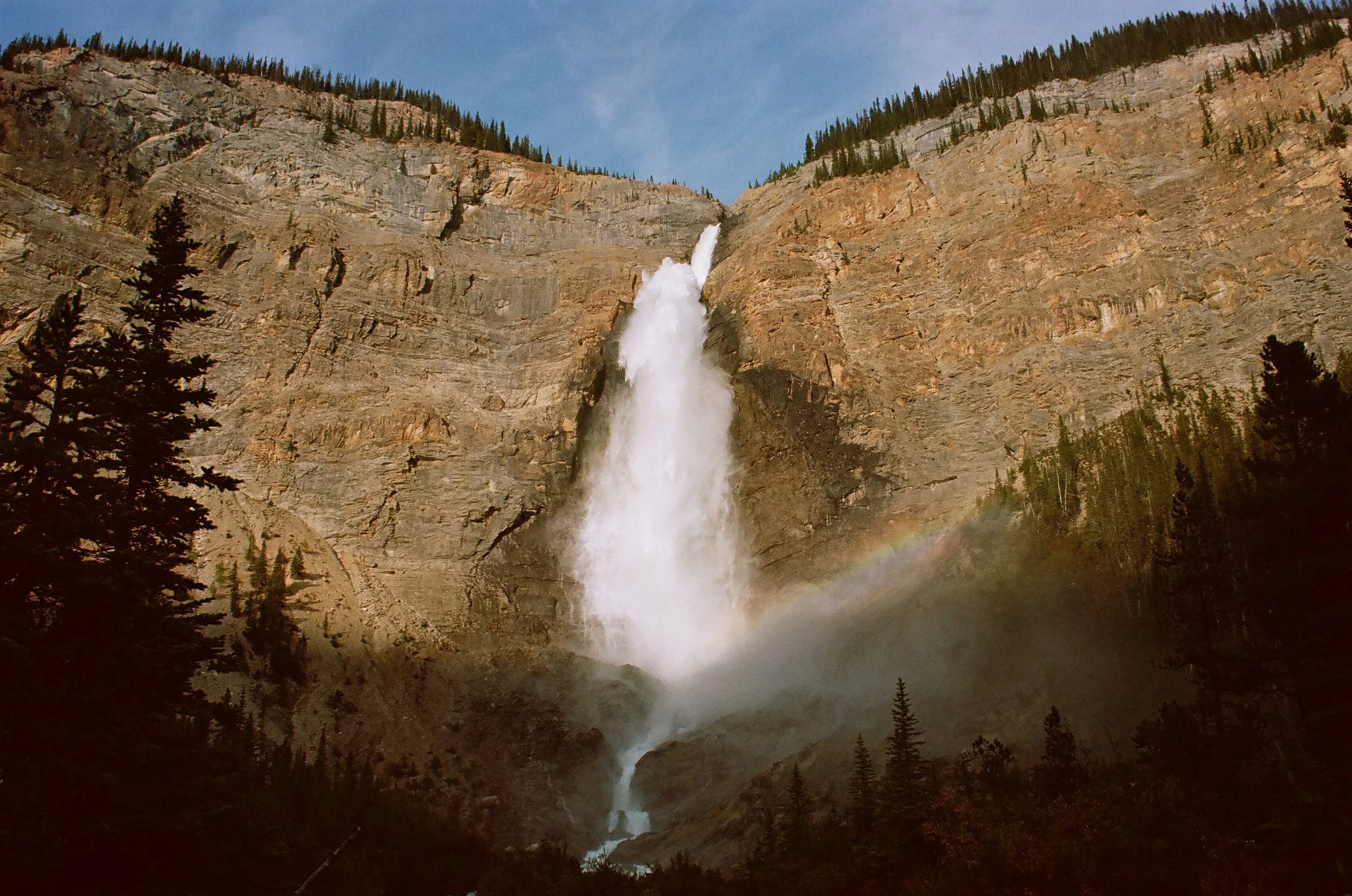 Dramatic waterfall cascading down a steep rocky mountain with trees and a rainbow near the base, under a blue sky.