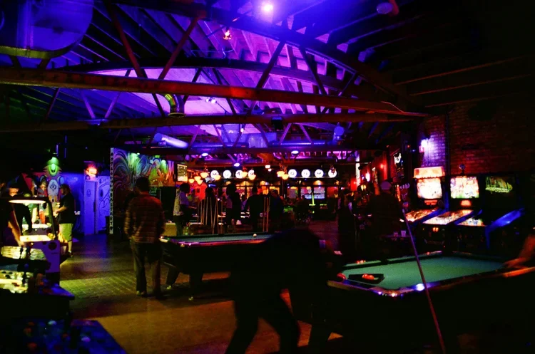 Indoor arcade with multiple pool tables, colorful neon lights, and people playing and socializing.