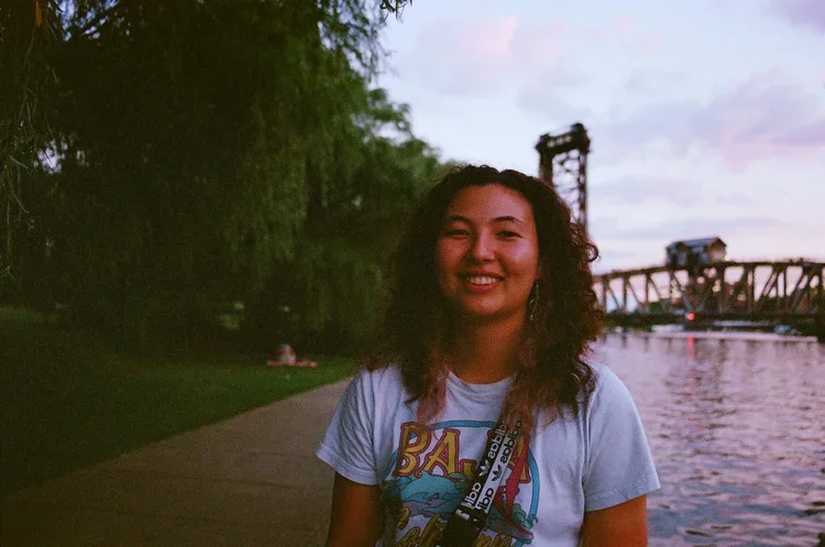 A smiling woman with curly hair standing near a river at sunset, with a bridge in the background.