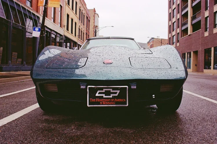 Black Chevrolet Corvette parked on an urban street with wet pavement and buildings on both sides.