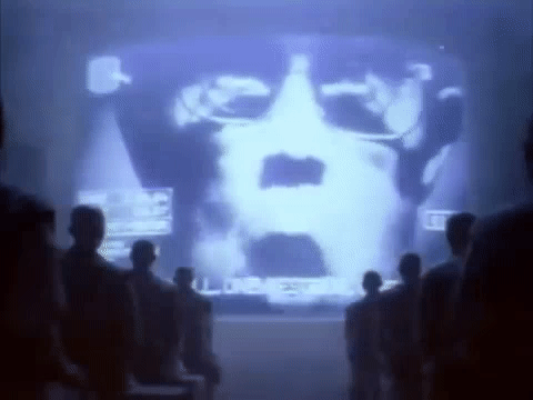 A large projection of a computer-generated face displayed on a screen with people seated in front of it, watching.
