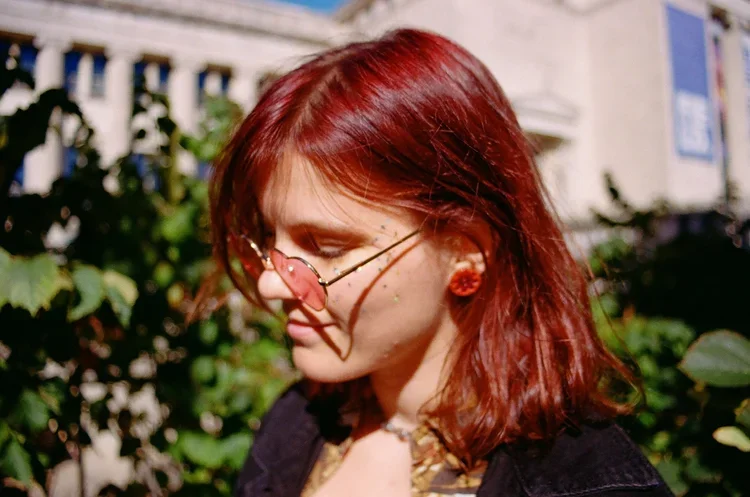 A woman with red hair, wearing round glasses with pink-tinted lenses, earrings, and a watch, standing outdoors in front of green plants and a building.