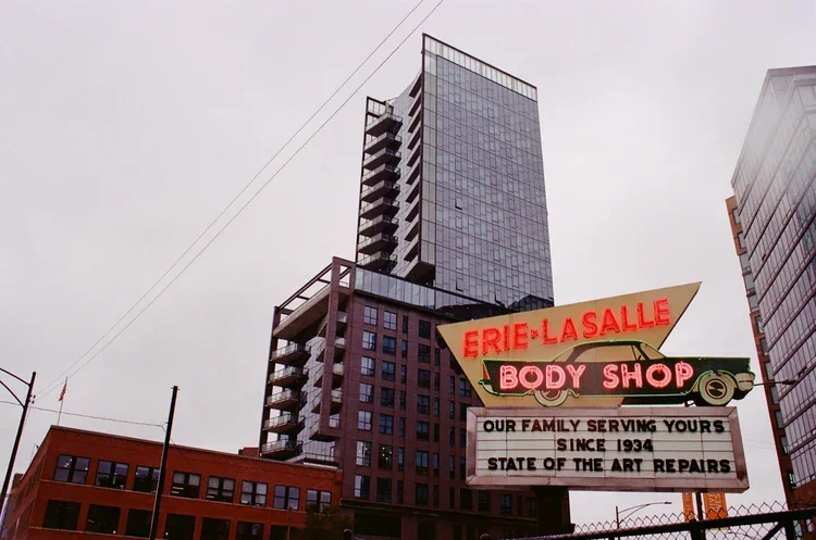 A tall modern high-rise building in a city with a neon sign for Erie-La Salle Body Shop in the foreground.
