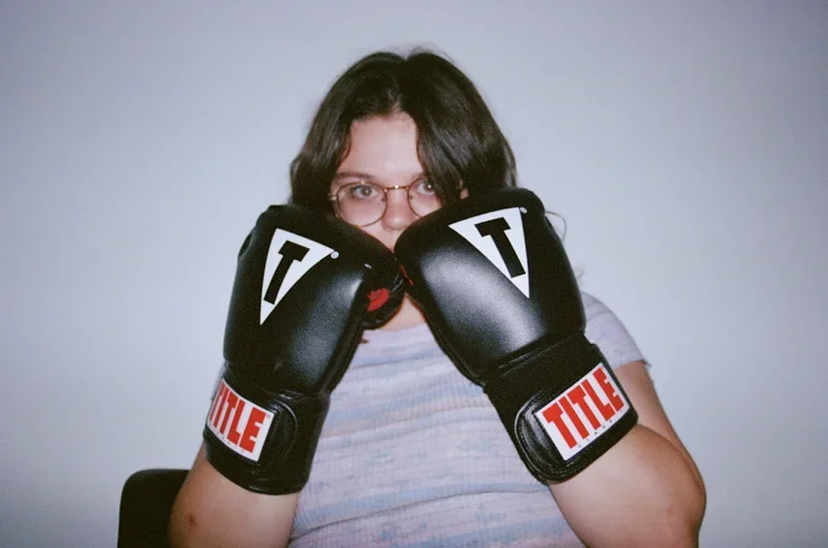 Person with glasses and dark hair wearing boxing gloves, standing against a plain background.