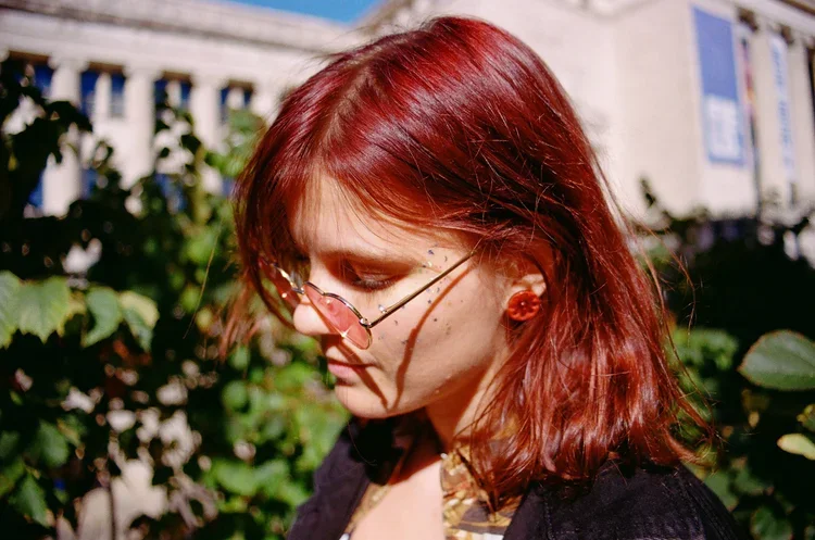A woman with shoulder-length red hair and glasses looking downward outdoors, with a building and green foliage in the background.