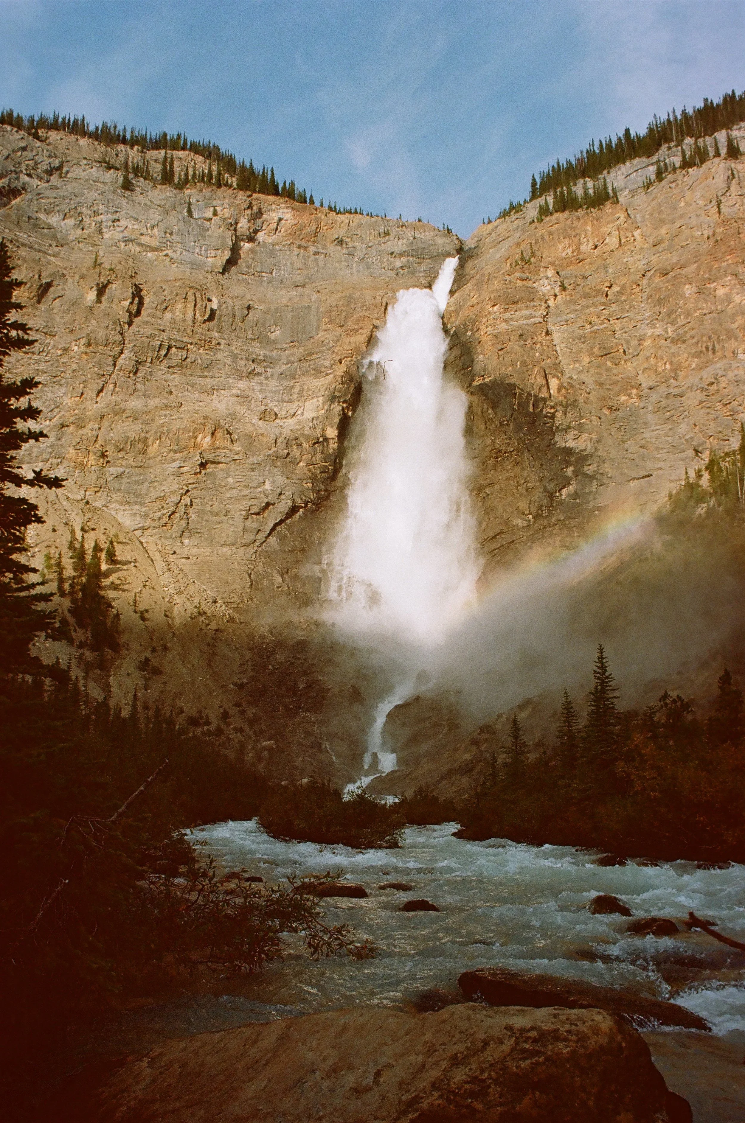 A waterfall cascading down a rocky cliff into a river below, with trees on the mountainside and a rainbow forming at the base of the waterfall.