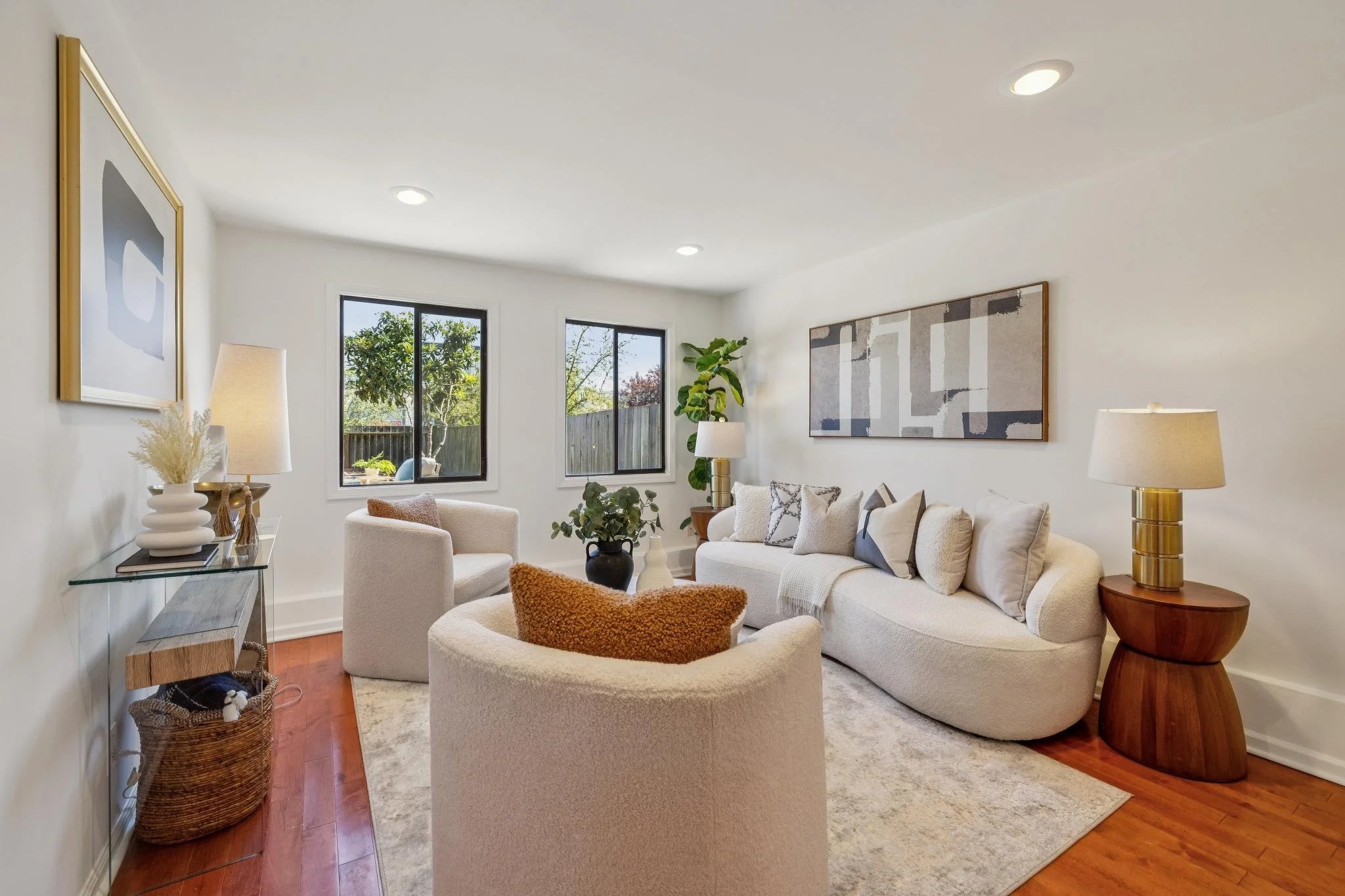 A bright, modern living room with a white sofa, two white armchairs, and wooden side tables with lamps. There are decorative pillows on the sofa, abstract artwork on the walls, and a large potted plant near the windows. The room has hardwood floors a