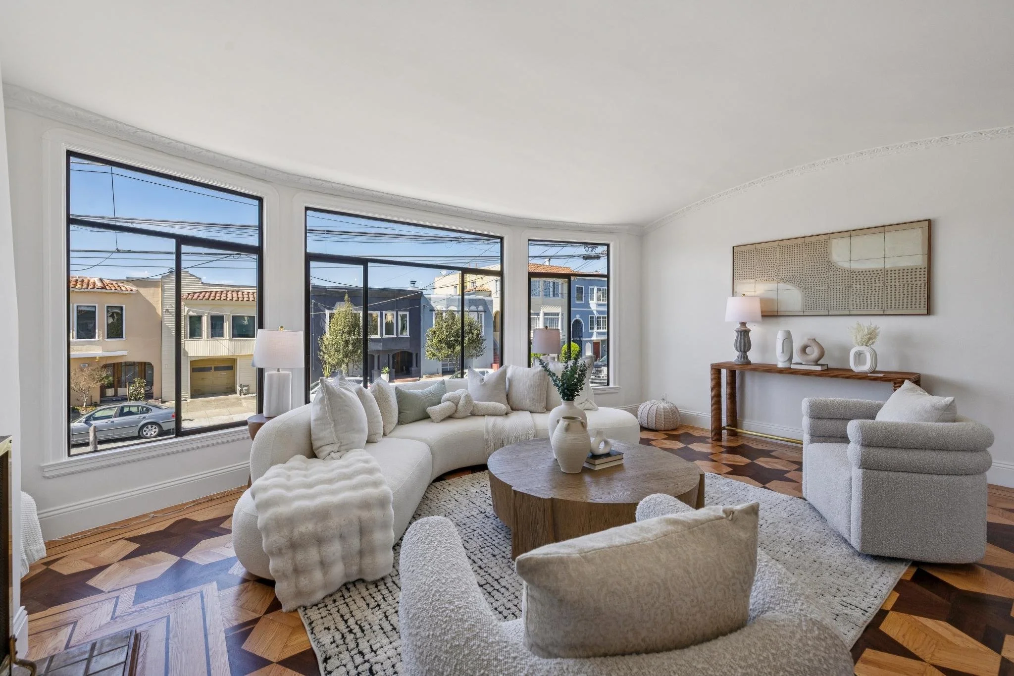 Bright living room with large windows, white curved sofa with pillows, modern armchair, round wooden coffee table with vases and books, and decorative art and vases on a wooden console against a white wall.