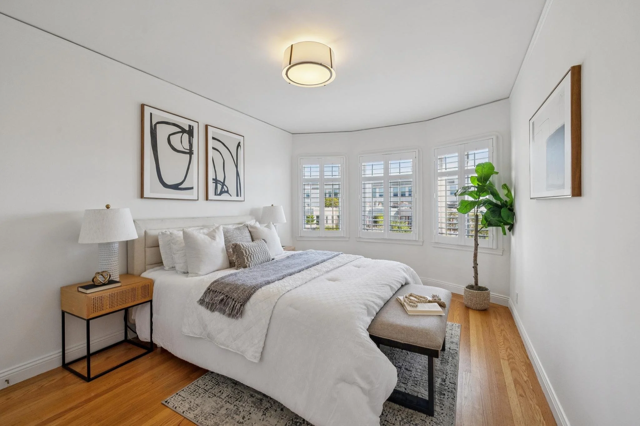 Bright bedroom with white walls, wooden floor, bed with white bedding, gray throw, and multiple pillows, large window with plantation shutters, potted plant, framed artwork, bedside table with lamp, and bench at foot of bed.