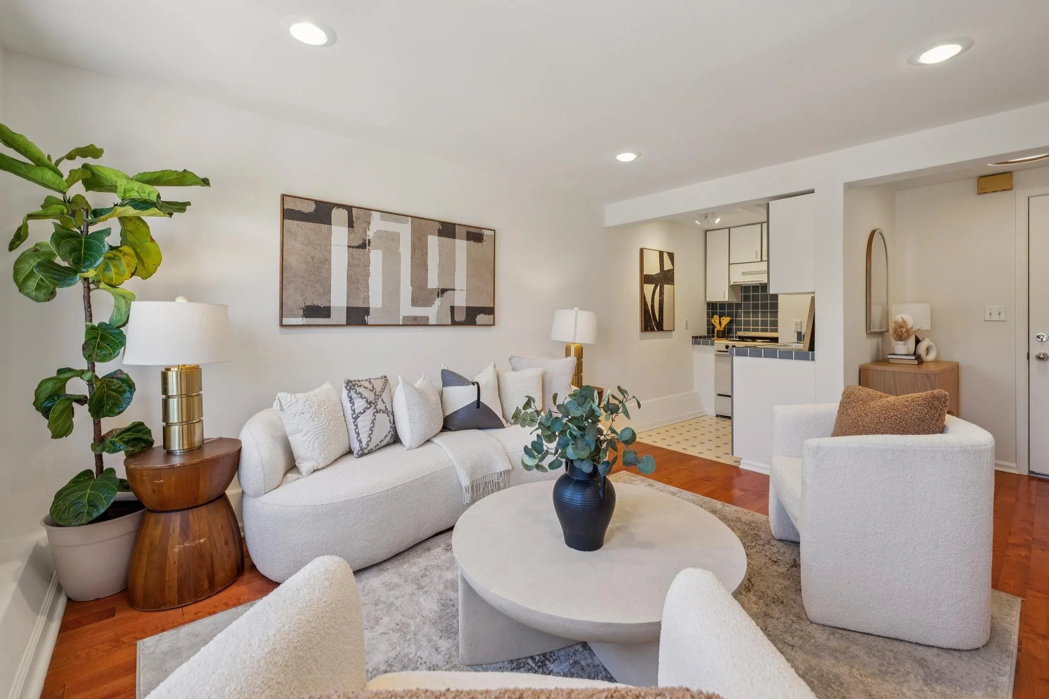Living room with white sofa, armchair, round coffee table with a black vase containing green foliage, large potted plant, modern artwork, floor lamps, and a view into the kitchen with white cabinets and black tiles.