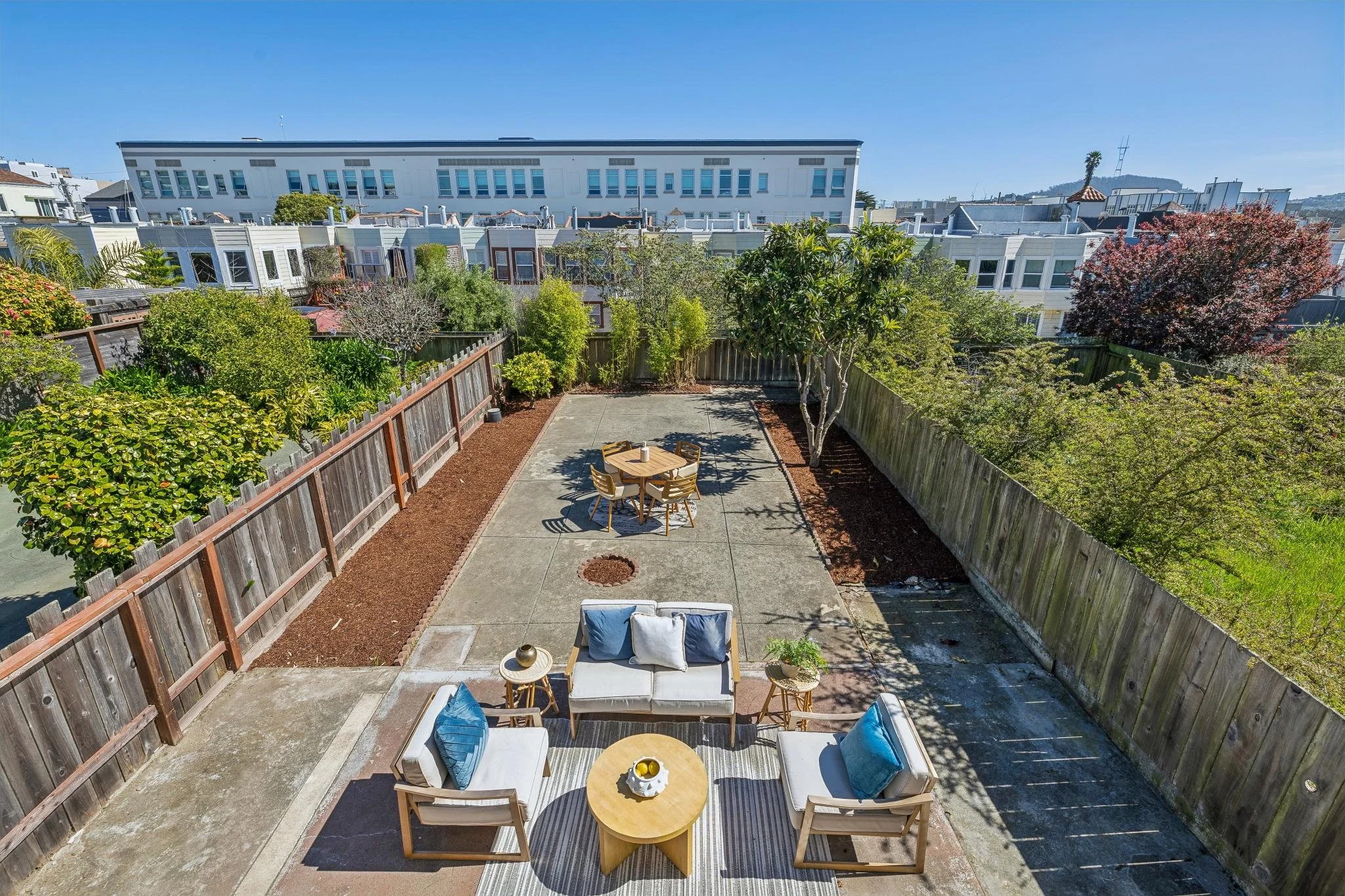 A backyard patio with outdoor furniture including a white sofa with blue and white pillows, two armchairs, a round coffee table with lemons, a small side table with a potted plant, and a separate dining table with chairs, surrounded by green trees an