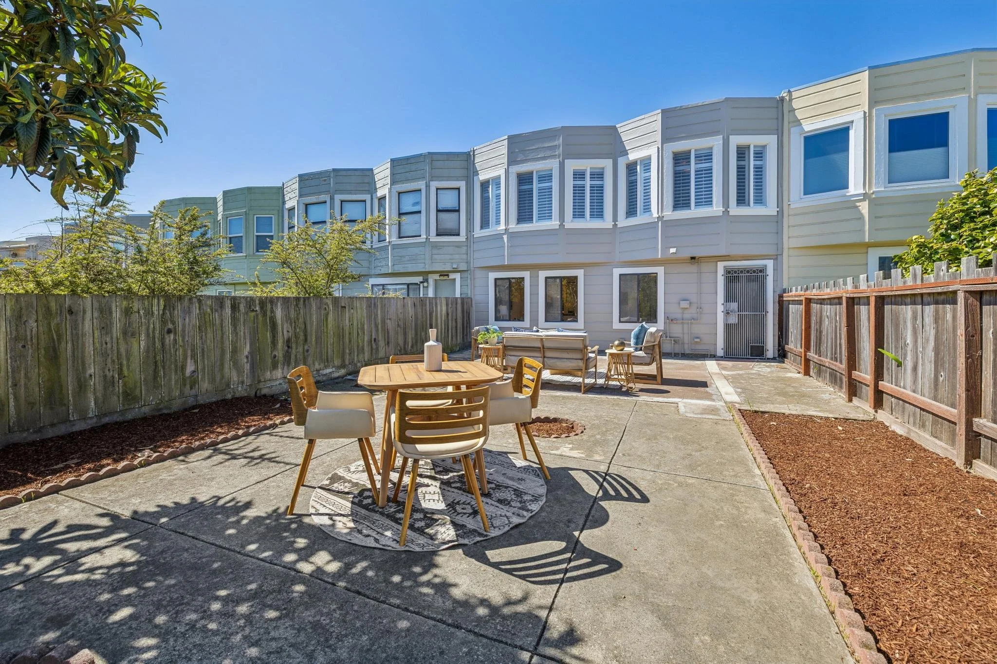 An outdoor patio with a round wooden table and six chairs on a circular rug, bordered by a wooden fence, with a row of colorful townhouses in the background under a clear blue sky.