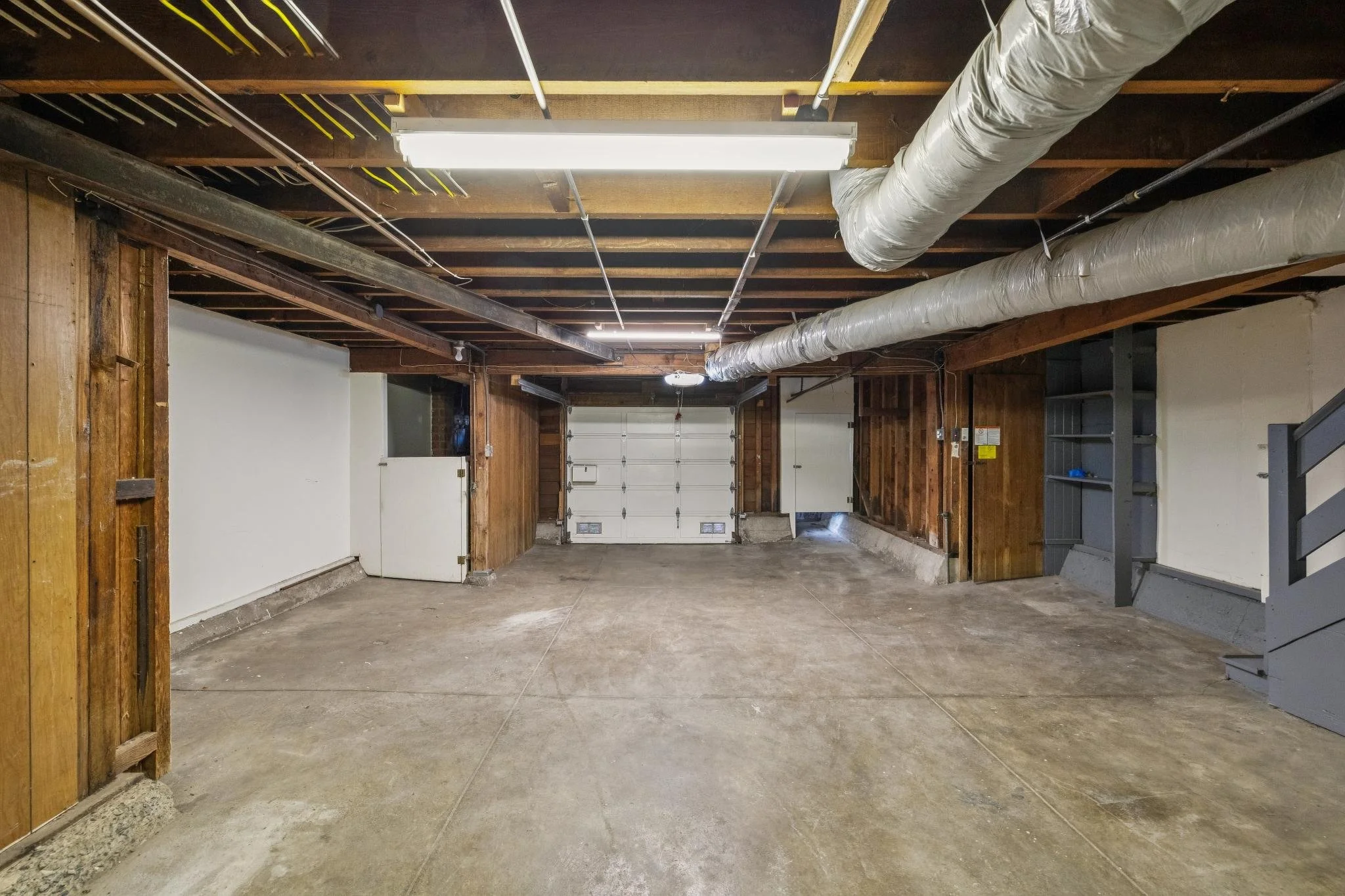 Empty garage with concrete floor, unfinished ceiling with exposed pipes and beams, and a white garage door at the back.