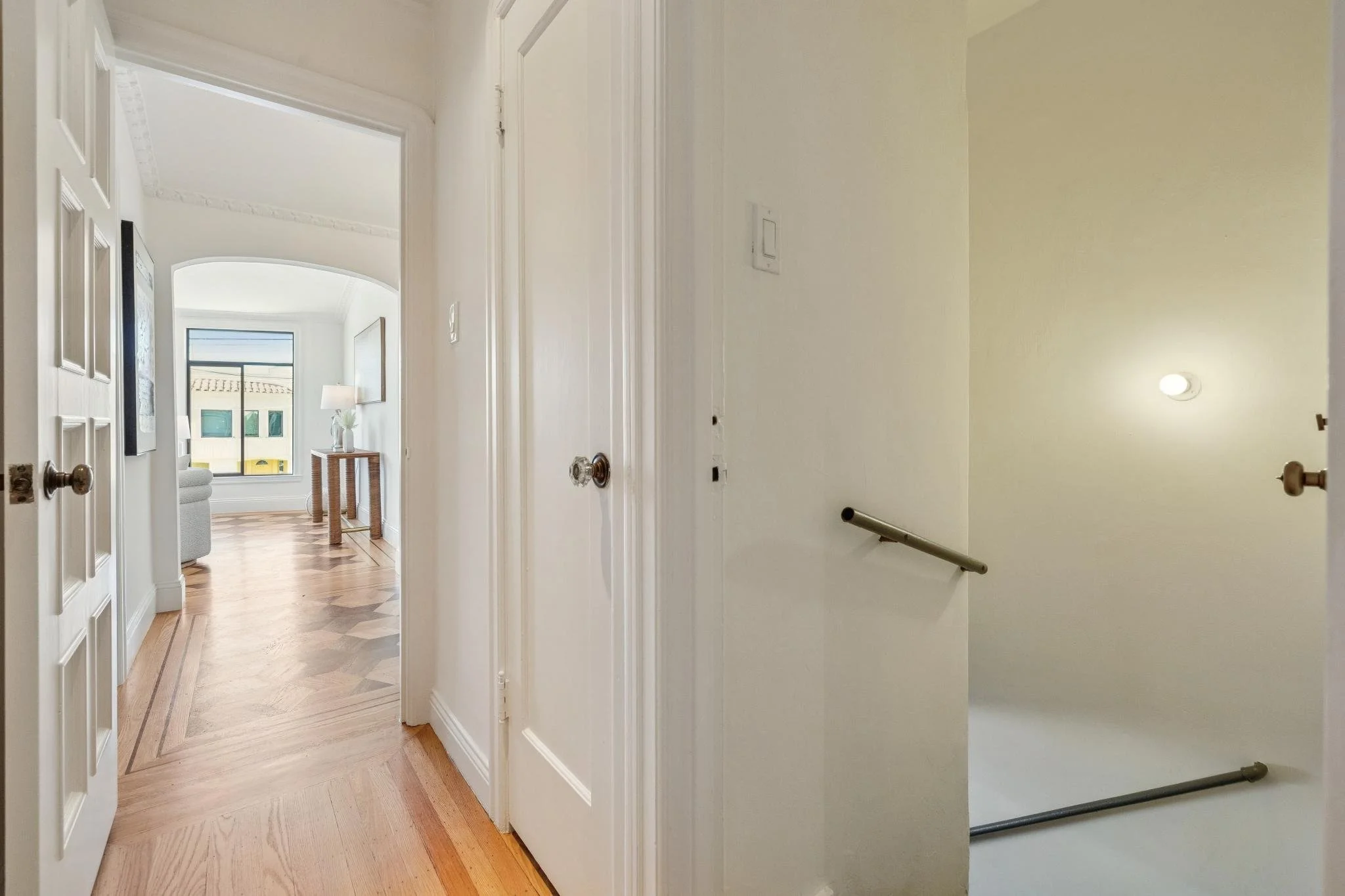 Interior view of a hallway leading to a living room with a large window, wood flooring, a side table with a lamp, and a gray armchair. The hallway has white walls, a staircase with a handrail, and a door on each side.