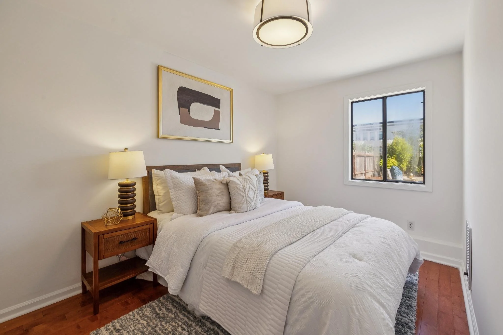 A cozy bedroom featuring a bed with white bedding and multiple pillows, two wooden nightstands with lamps, an abstract wall art piece, a window with outdoor views, and a ceiling light fixture.
