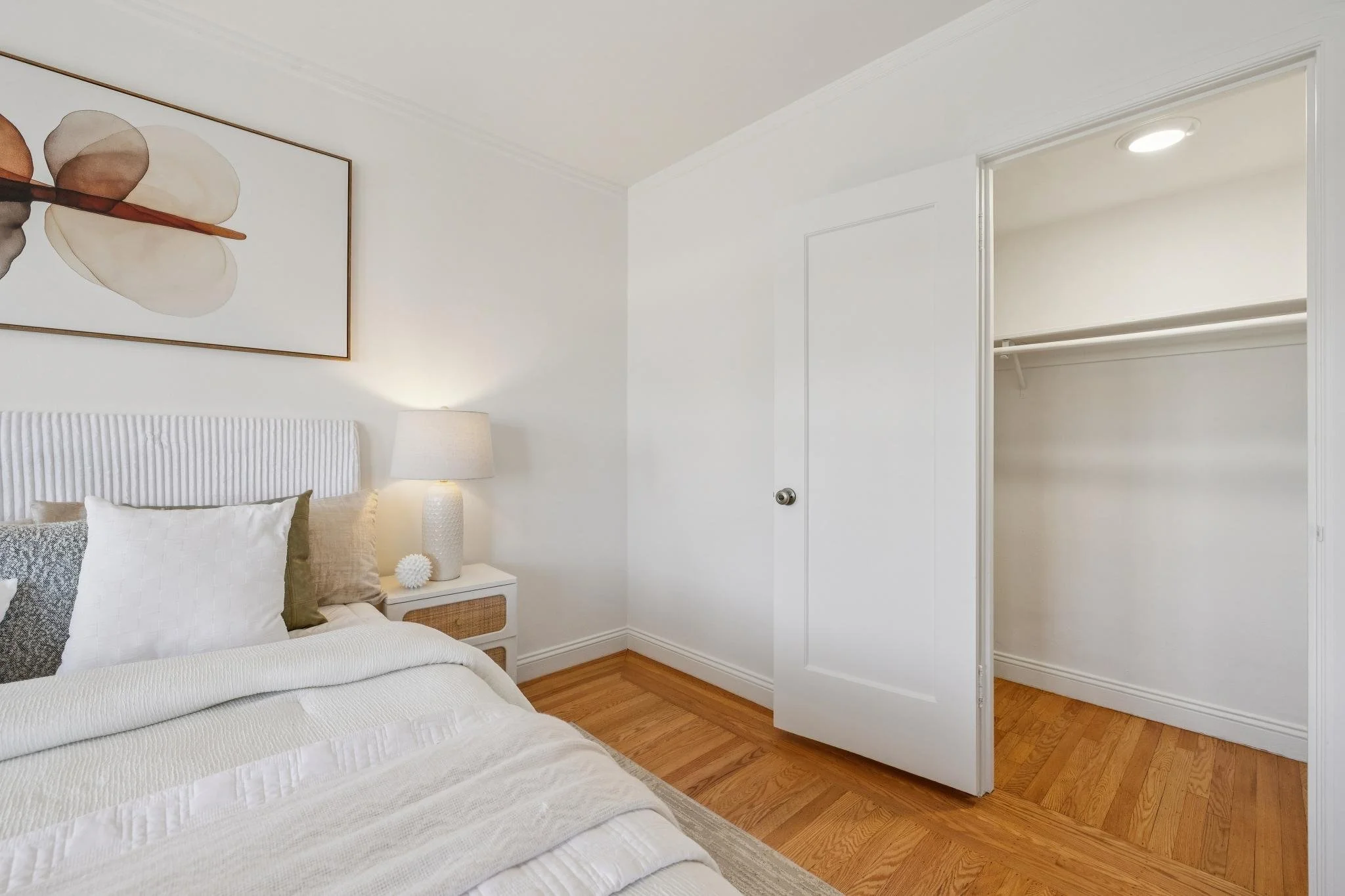 Bedroom with white walls, a bed with white bedding and pillows, a nightstand with a lamp, and an empty closet with a door open.