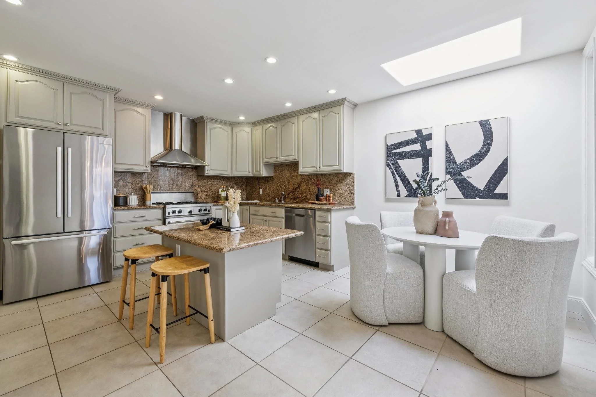 Modern kitchen with beige cabinets, granite countertops, stainless steel appliances, a small island with two stools, and a dining area with a round table and four chairs. Decor includes vases and abstract artwork. Natural light from a skylight illumi
