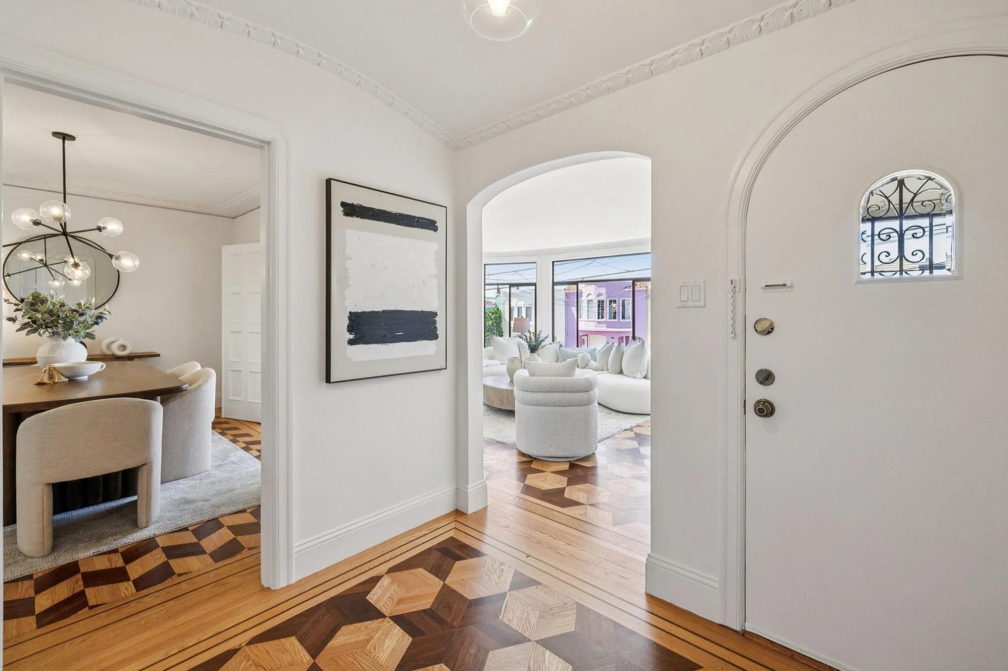 View of a bright, modern living room with large windows, white furniture, and a purple house visible outside, seen through an archway from an entryway with a decorative wooden floor.