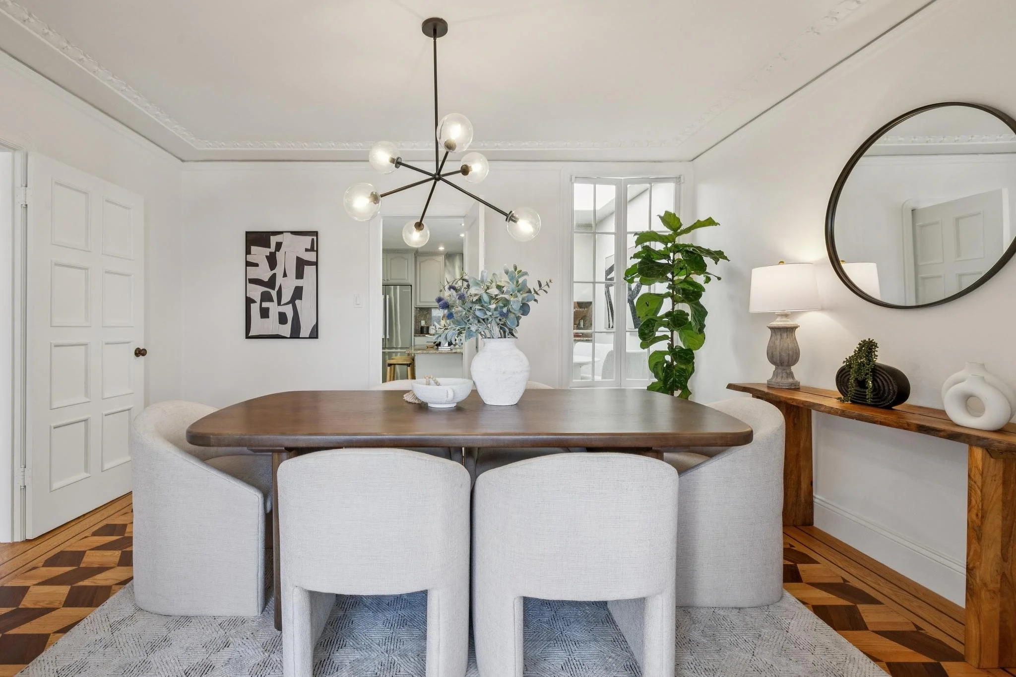 Dining room with a wooden oval table, six light-colored upholstered chairs, a modern chandelier with multiple globes, a wall mirror, a table lamp, potted plant, and decorative vases and artwork on a console table.