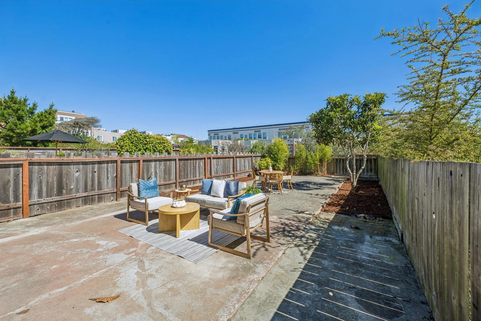 Outdoor patio area with seating, including a white couch and chairs with cushions, a small wooden table, and a dining table with chairs, surrounded by a wooden fence and trees, under a clear blue sky.