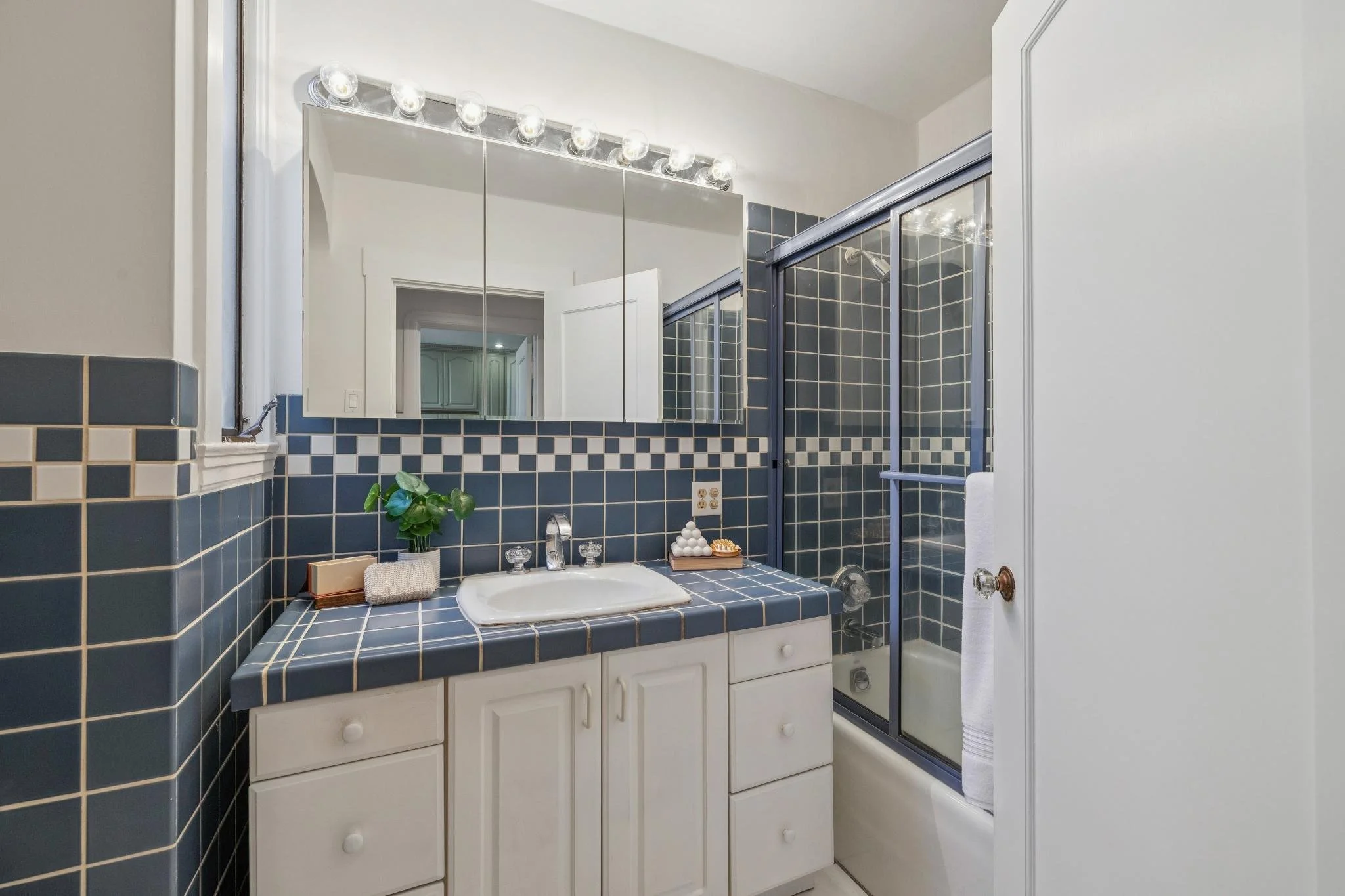 A small bathroom with a blue tiled sink and backsplash, a white cabinet, a mirror above the sink, and a shower with glass sliding doors. There is a small plant and toiletries on the sink counter, and a towel hanging on the shower door handle.
