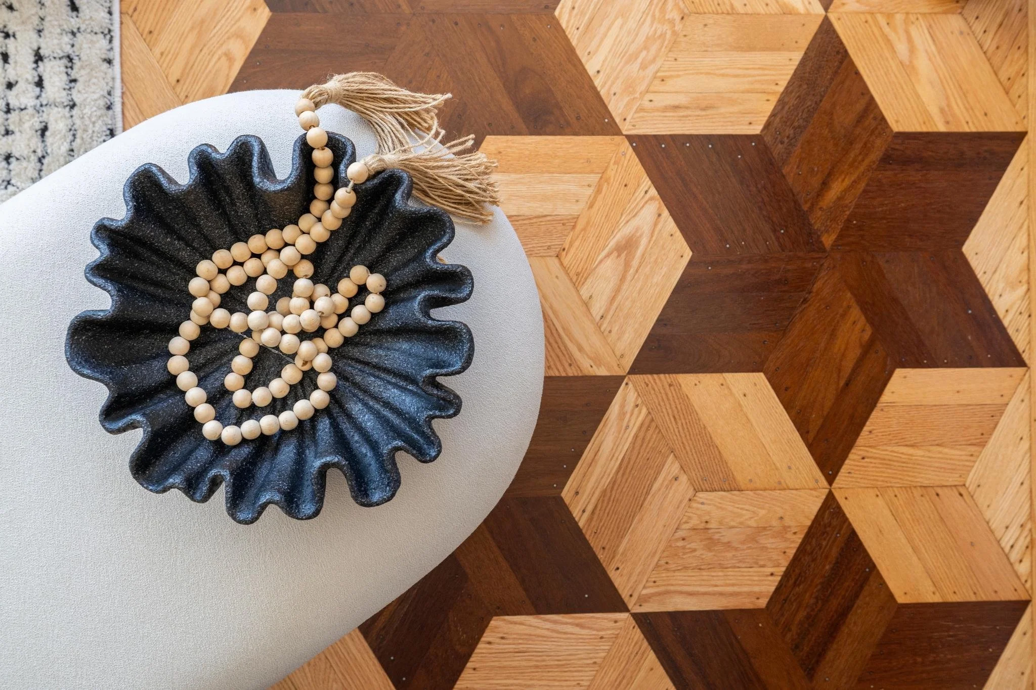 A black textured bowl with a scalloped edge holding a string of light-colored wooden beads with some tassels, placed on a beige surface next to a geometric patterned wooden floor.