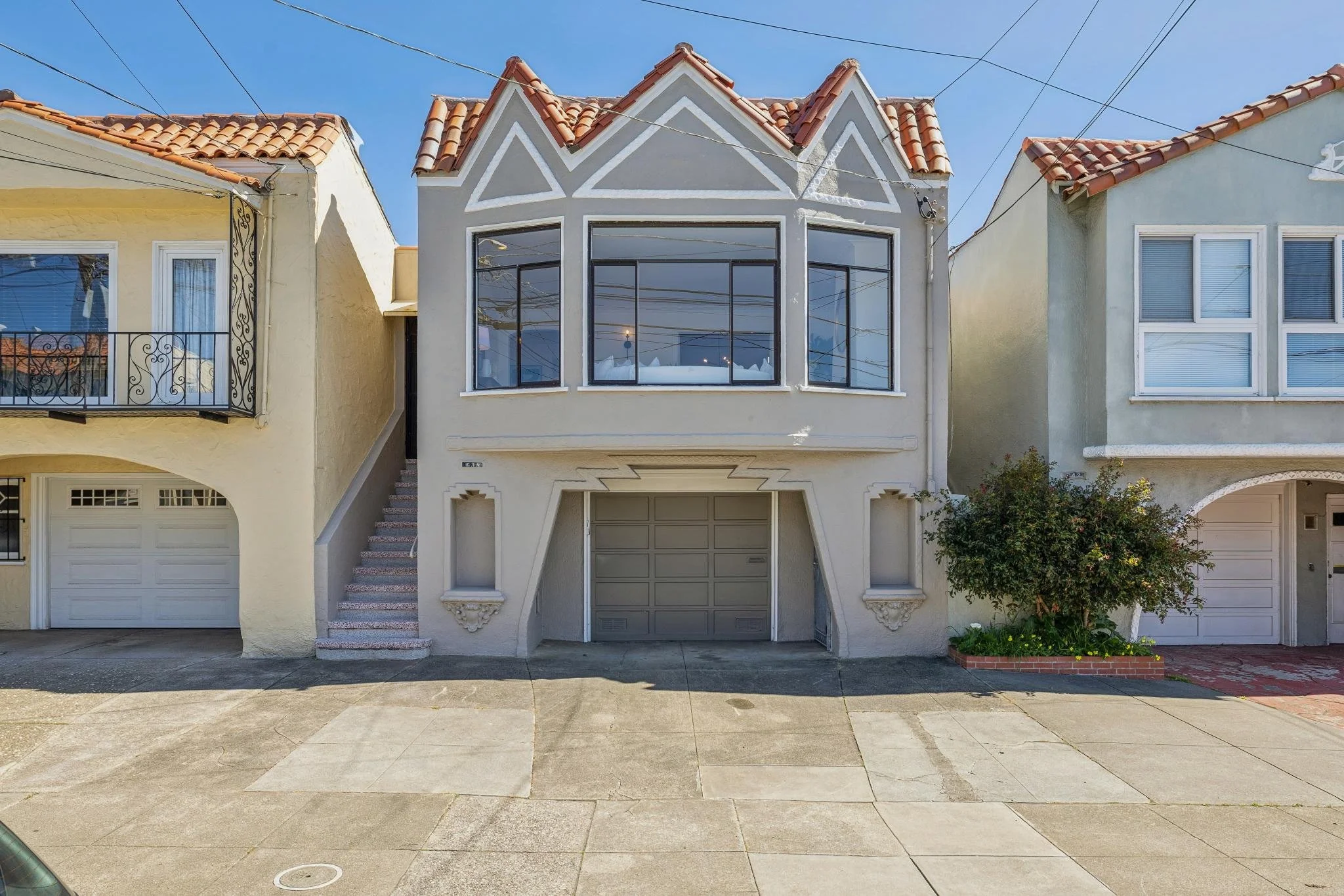 A three-story house with a unique architectural facade, featuring a garage on the ground level, large windows on the second floor, and a decorative gabled roof with red tiles. The house is painted in light colors and is flanked by neighboring homes on a sunny day.