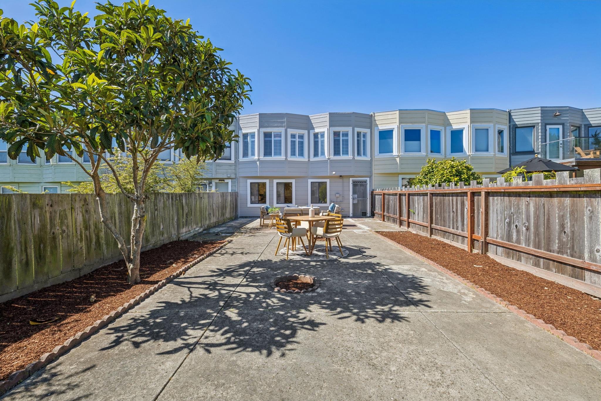 Backyard patio with outdoor dining table and chairs, surrounded by wooden fences, a tree providing shade, and a row of townhouses in the background under a clear blue sky.