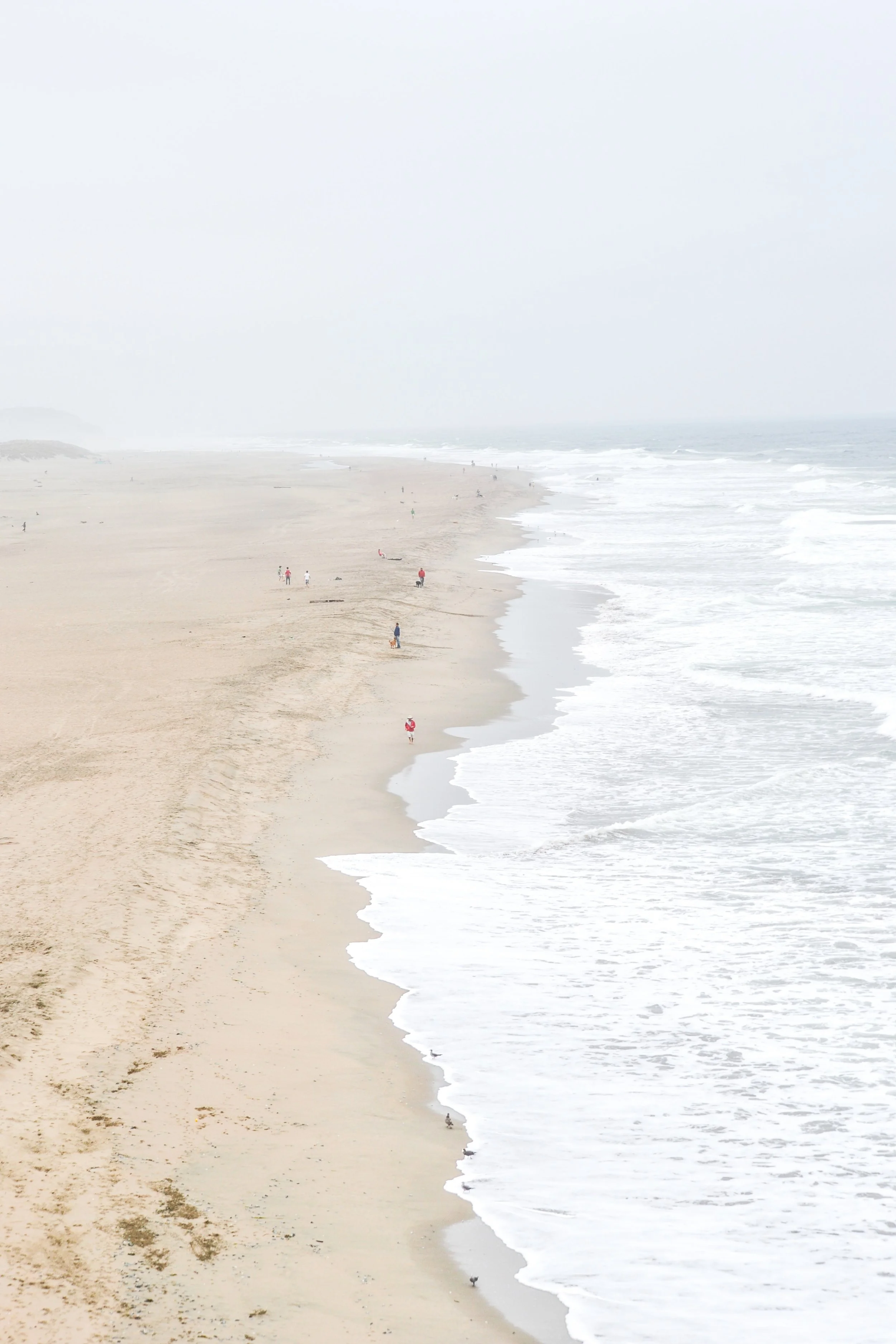 A misty beach with a few people walking along the shore, waves gently crashing onto the sand, and distant dunes in the background.