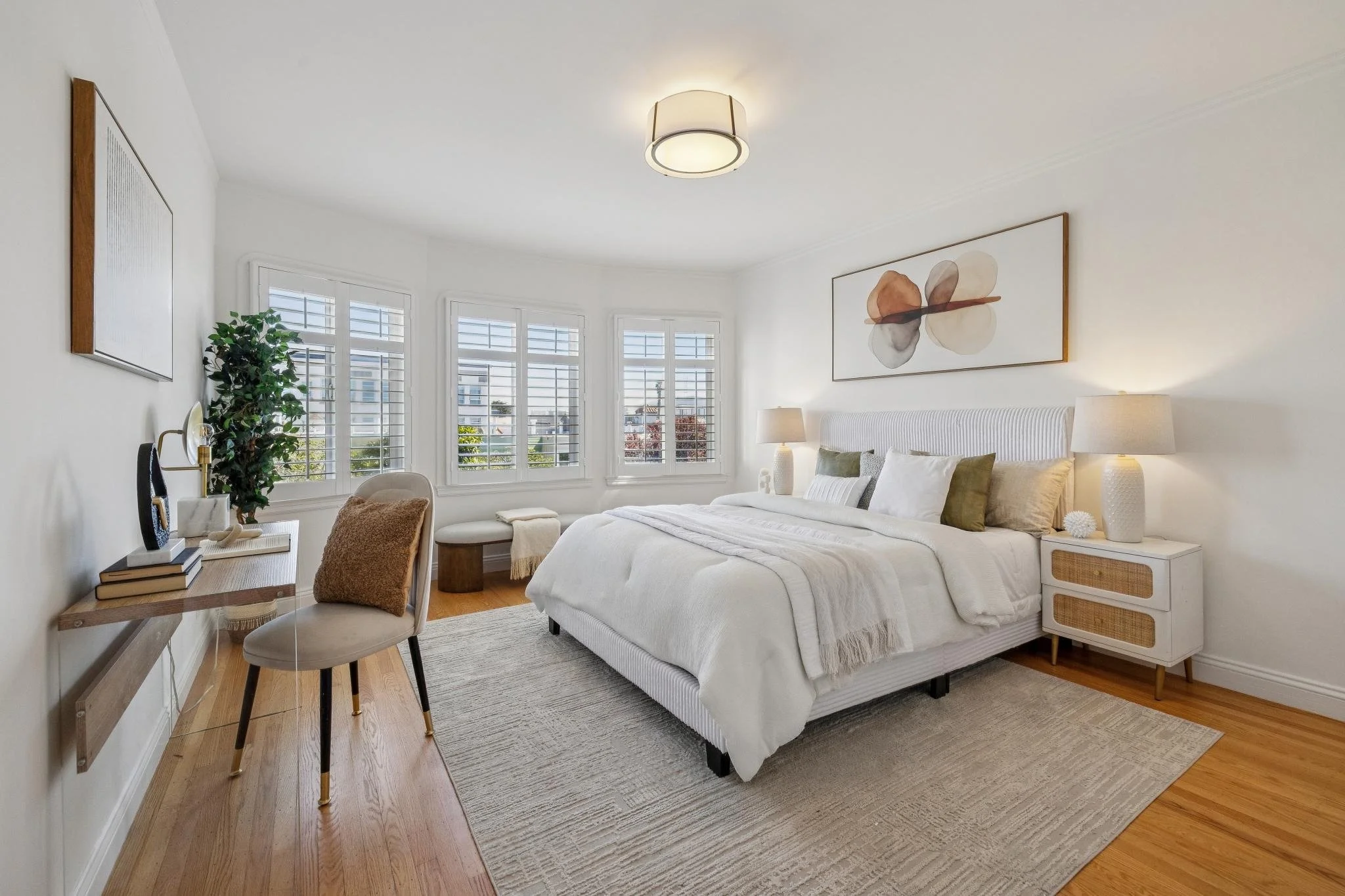 A bright bedroom with white walls, a white bed with multiple pillows, two white bedside lamps, a large abstract artwork above the bed, a window with white shutters, a chair with a pillow, and a rug on hardwood floor.