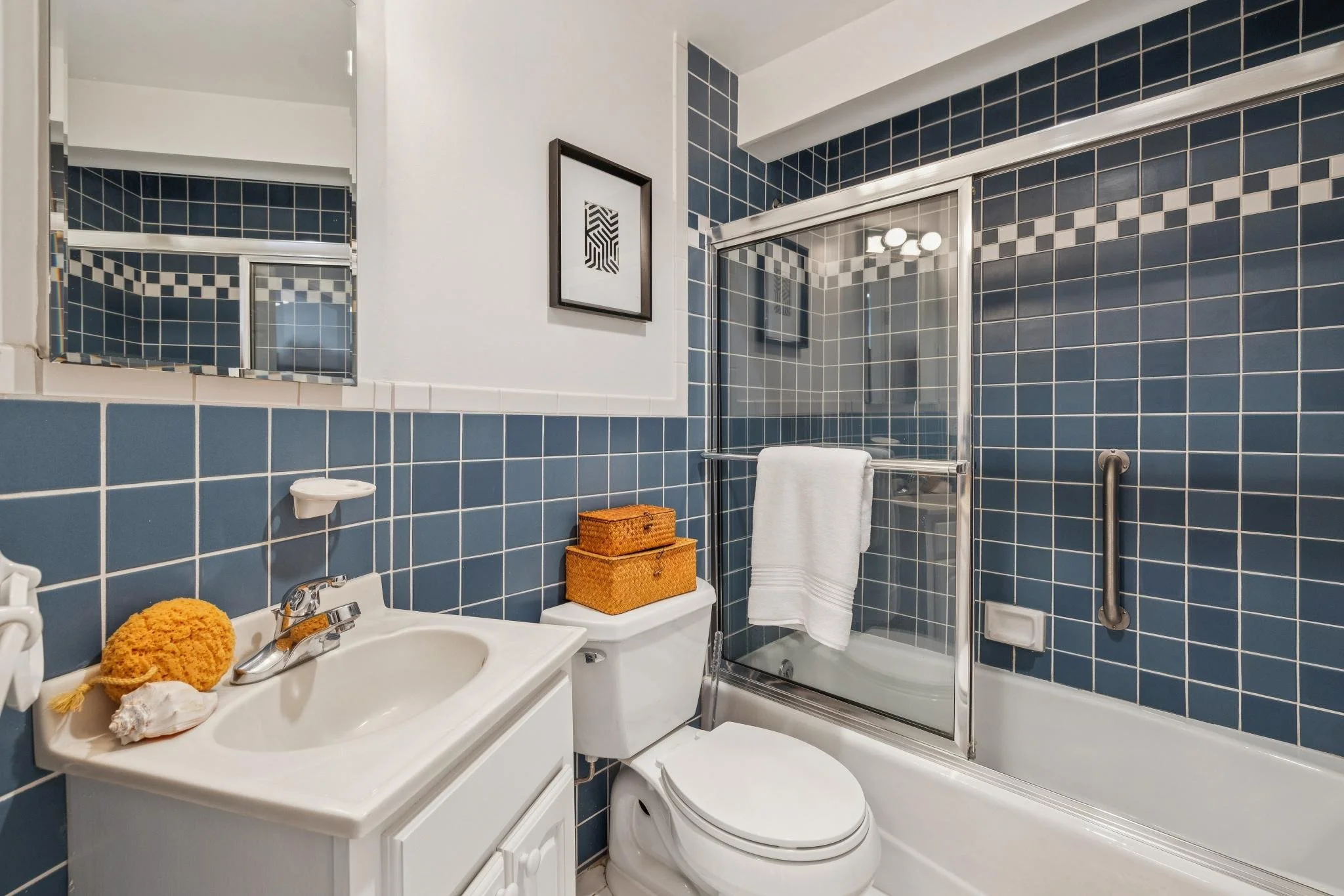 Bathroom with blue tiled walls, a white sink with a yellow loofah and shells, a toilet with two orange baskets on top, a mirror, and a shower with a sliding glass door and white towel.