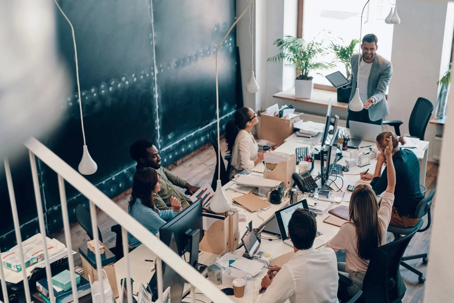 Office meeting with diverse group of professionals seated around a large desk, with a man standing and presenting, in a bright room with large windows, plants, and multiple computer monitors.